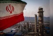 Iranian flag waving in the foreground with an oil refinery and industrial plant in the background.