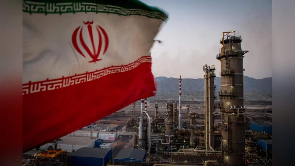 Iranian flag waving in the foreground with an oil refinery and industrial plant in the background.
