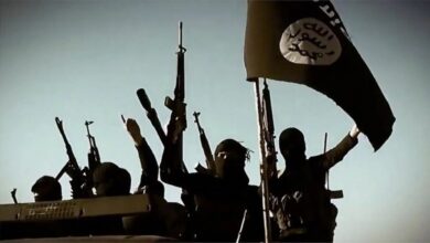 An image of armed militants, wearing black clothing and balaclavas, holding rifles in a desert landscape. The militants are shown in military formation, with a black flag featuring Arabic text in the background.