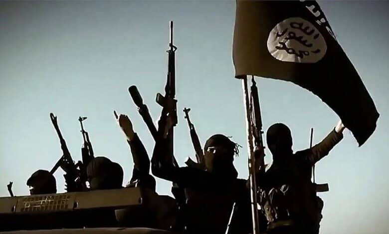 An image of armed militants, wearing black clothing and balaclavas, holding rifles in a desert landscape. The militants are shown in military formation, with a black flag featuring Arabic text in the background.