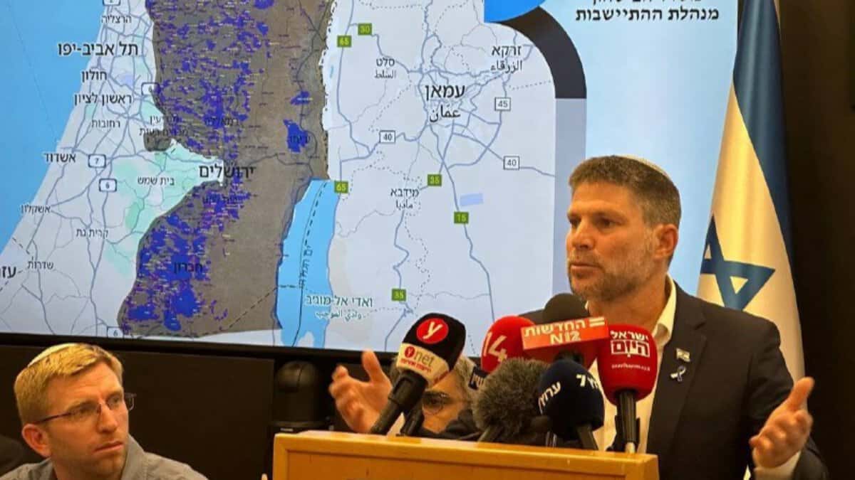 Bezalel Smotrich speaking at a press conference with a map of the West Bank behind him.