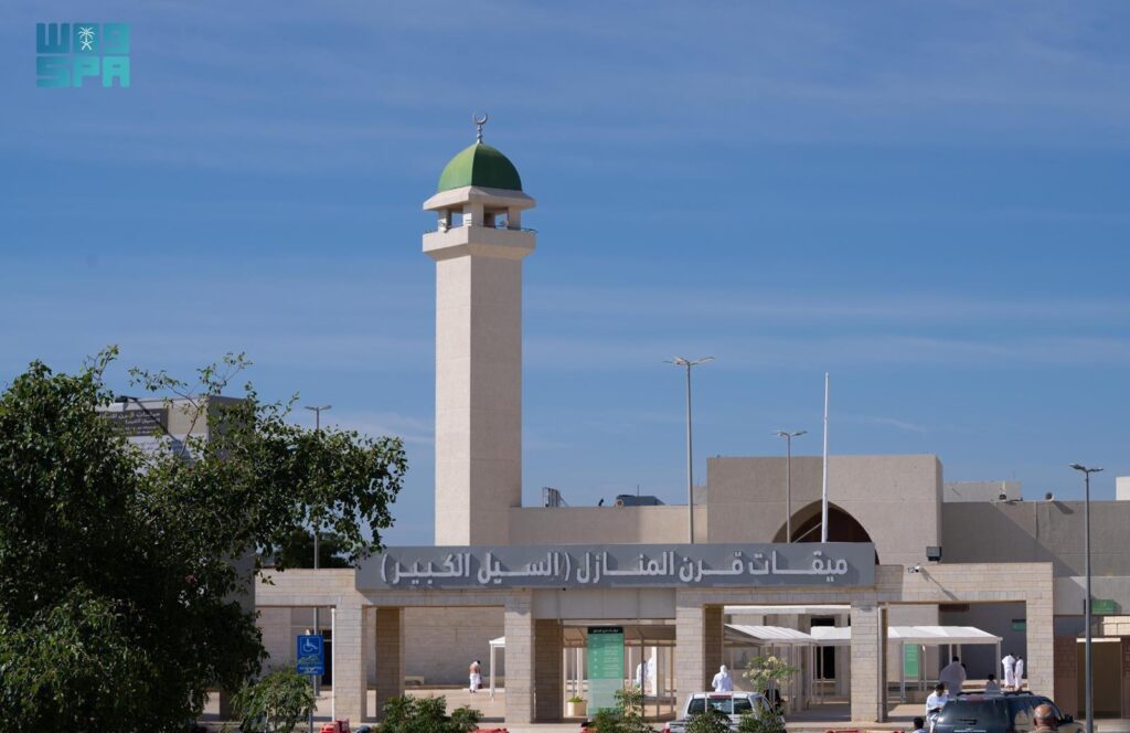 A close-up view of the entrance to Miqat Qarn Al-Manazil (Al-Sail Al-Kabeer) mosque, featuring the sign in Arabic, a minaret with a green dome, and pilgrims entering the premises.