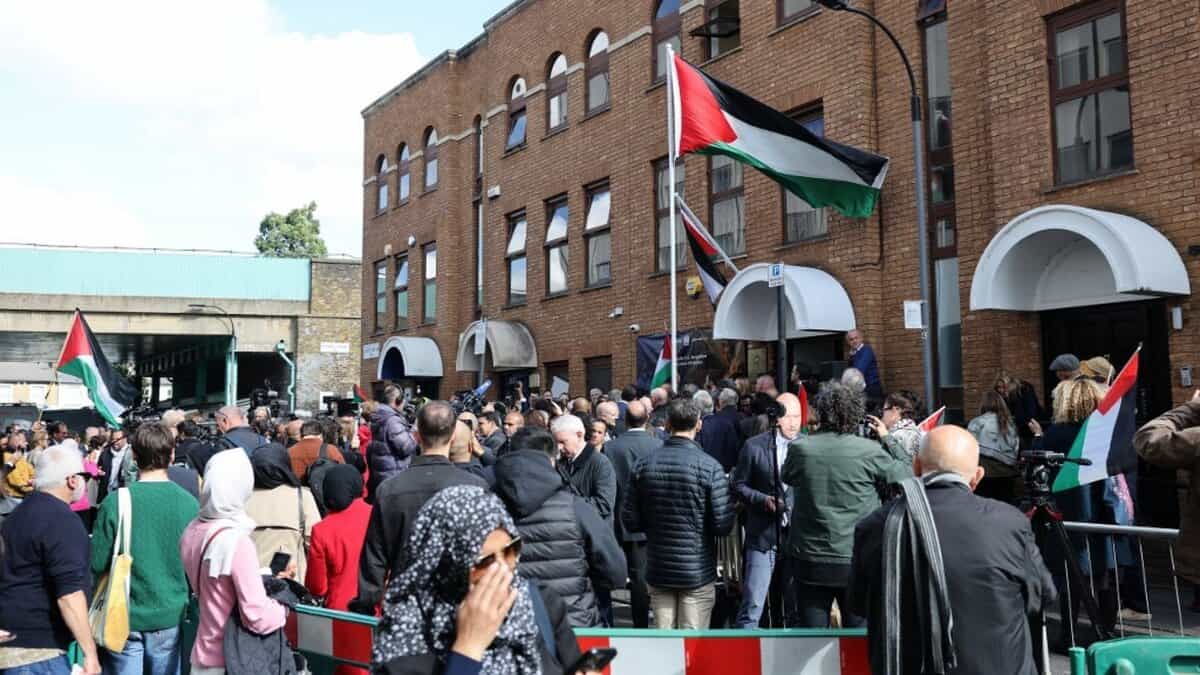 A diverse crowd of men and women, many wearing headscarves and traditional attire, gathers outside a brick building adorned with Palestinian flags during a flag-raising ceremony marking the UK's recognition of a Palestinian state, London, September 22, 2025.
