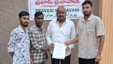Four men standing before a "Pravasi Prajavani" banner with political figures and the Telangana logo, one holding a document, dressed in casual and formal attire.