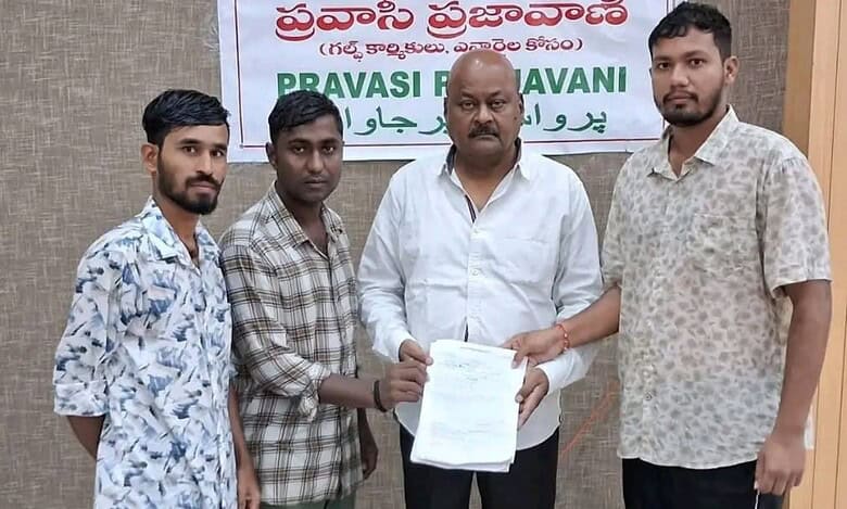 Four men standing before a "Pravasi Prajavani" banner with political figures and the Telangana logo, one holding a document, dressed in casual and formal attire.
