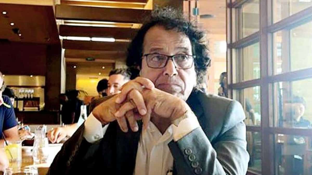 A seated older man with glasses and curly hair rests his hands together at a table in a well-lit café or restaurant.