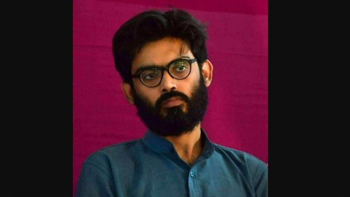Young man with glasses and beard, wearing a blue shirt, posing against a pink background.