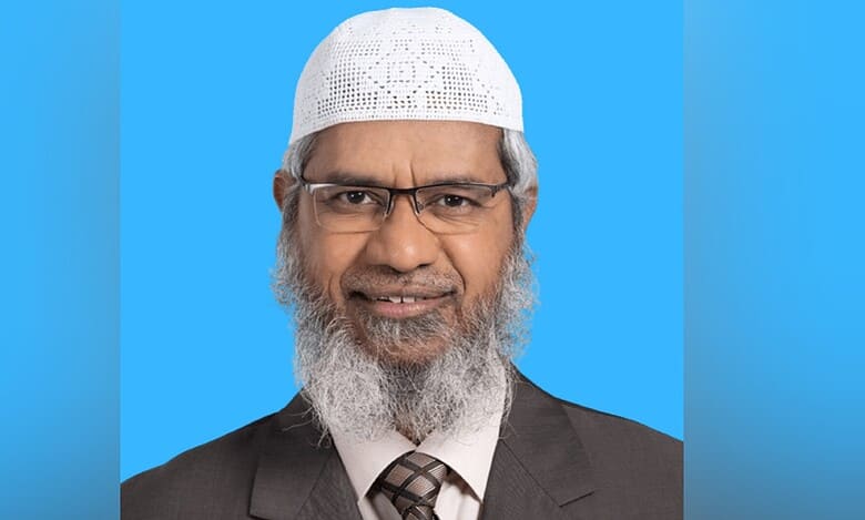 Zakir Naik pictured in a suit and white cap against a blue background.
