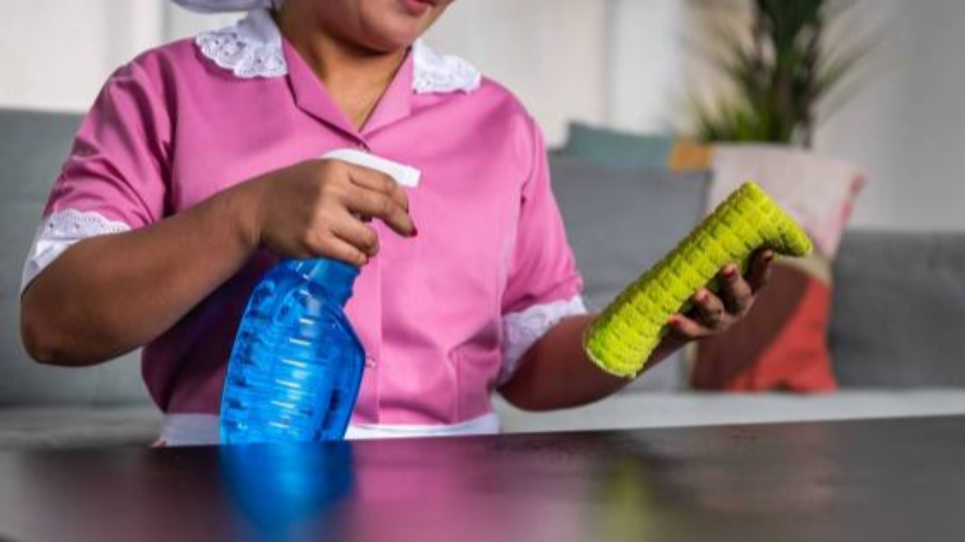 A domestic worker in uniform cleaning a table with a spray bottle and cloth.
