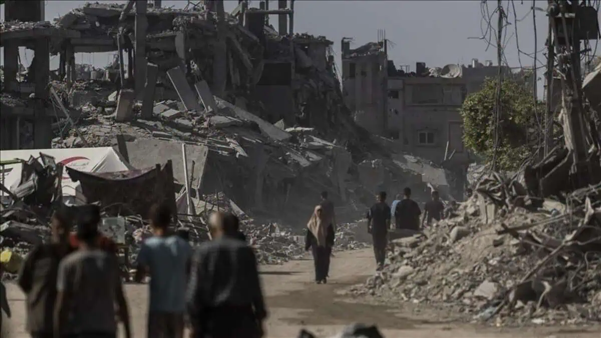 People walk through a street lined with destroyed buildings in a war-torn area.