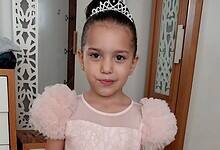 A young Palestinian girl, Hind Rajab, wearing a pink dress and tiara, smiles softly while standing indoors.