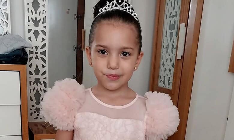 A young Palestinian girl, Hind Rajab, wearing a pink dress and tiara, smiles softly while standing indoors.