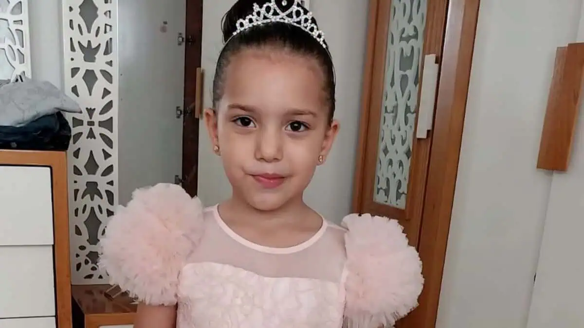 A young Palestinian girl, Hind Rajab, wearing a pink dress and tiara, smiles softly while standing indoors.