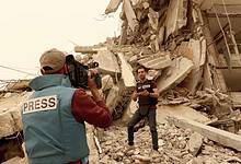 Two journalists wearing press vests report amid the rubble of a destroyed building in Gaza.