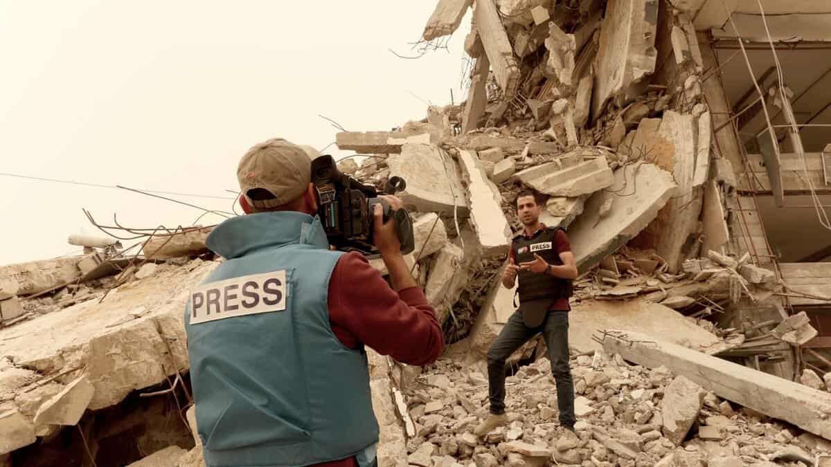 Two journalists wearing press vests report amid the rubble of a destroyed building in Gaza.