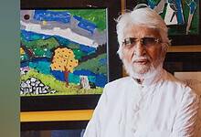 Indian painter MF Husain, dressed in a white kurta with a beard and white hair, wearing sunglasses, standing in front of colourful modern artworks.