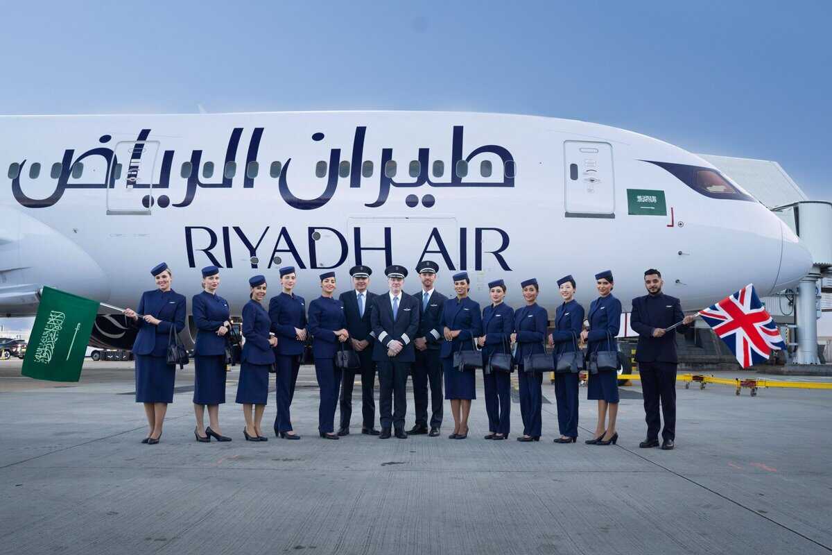 Riyadh Air flight crew and pilots pose in front of a Boeing 787 aircraft at Heathrow Airport, holding Saudi and British flags.