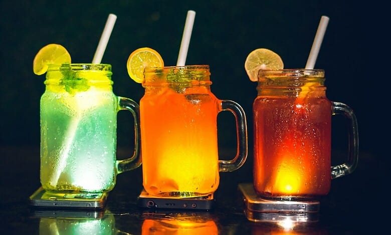 Three colourful sugar-sweetened drinks in glass jars with lemon slices and straws placed on a dark surface.