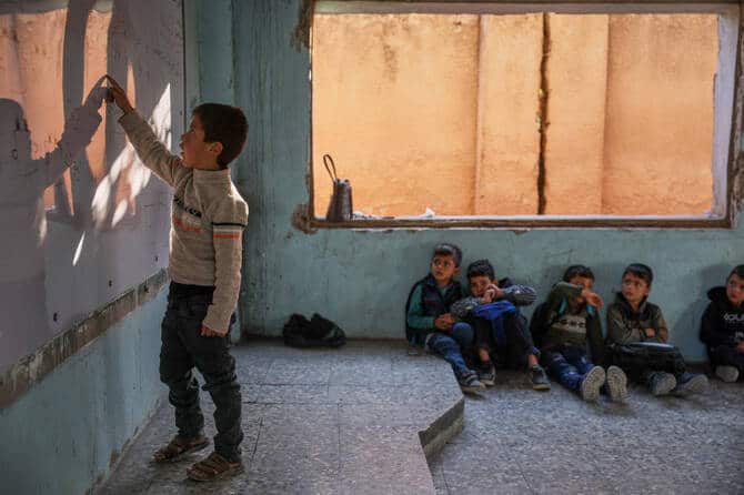 A Syrian boy writes on a damaged classroom board as other children sit on the floor in Idlib.