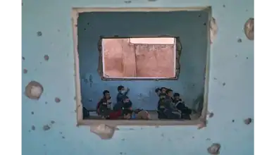 Children sit on the floor of a bullet-riddled classroom in Idlib, Syria, seen through a damaged wall.