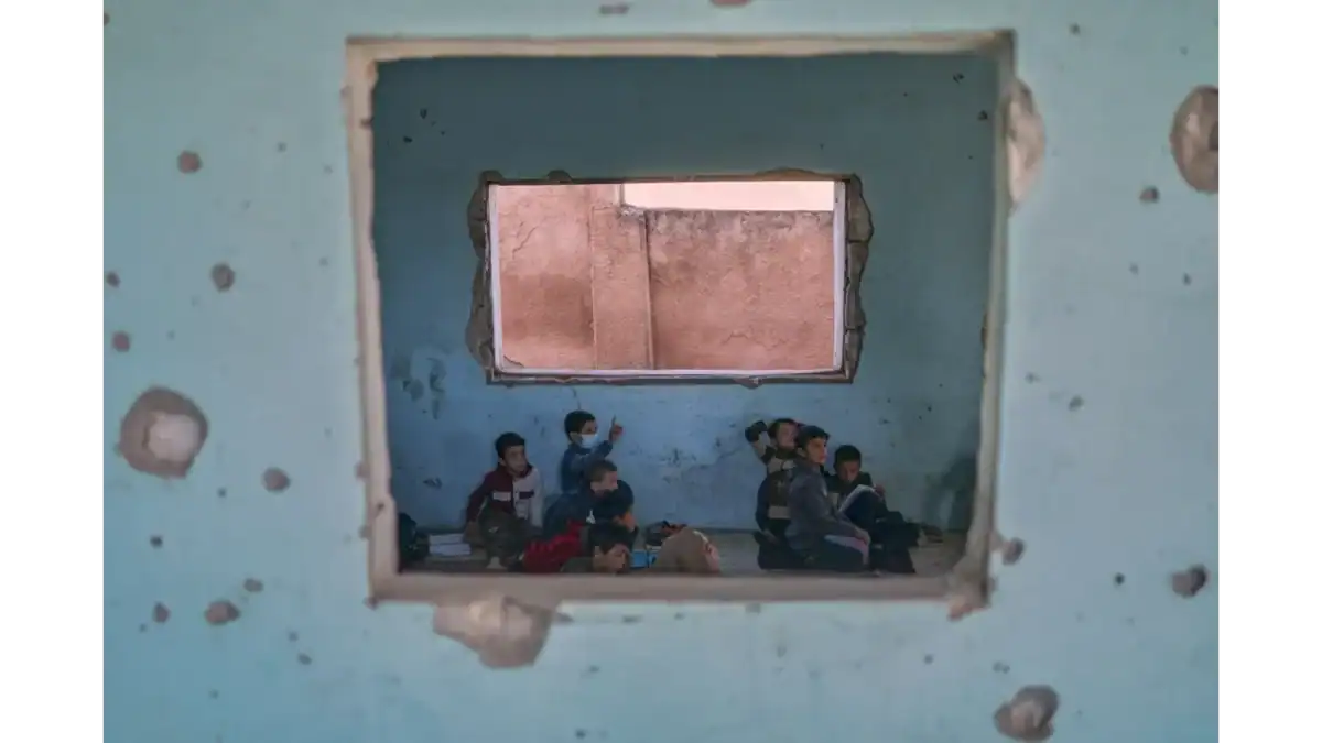 Children sit on the floor of a bullet-riddled classroom in Idlib, Syria, seen through a damaged wall.