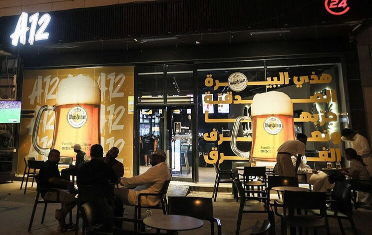 Night view of A12 Cafe in Riyadh with customers seated outside and large posters advertising Warsteiner 0.0 percent alcohol-free beer on the storefront.