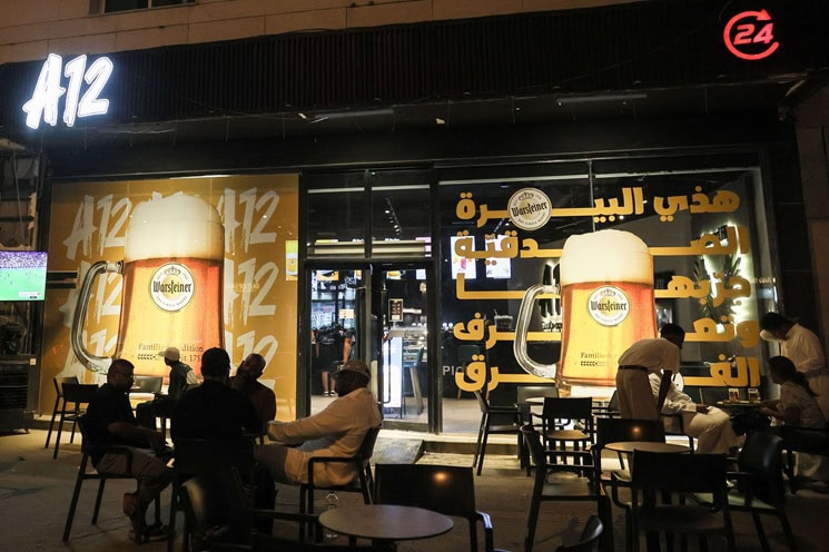 Night view of A12 Cafe in Riyadh with customers seated outside and large posters advertising Warsteiner 0.0 percent alcohol-free beer on the storefront.