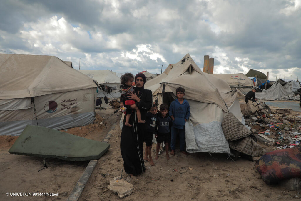 A displaced mother with her children stands outside a makeshift tent in Gaza as cold weather and rain worsen living conditions.