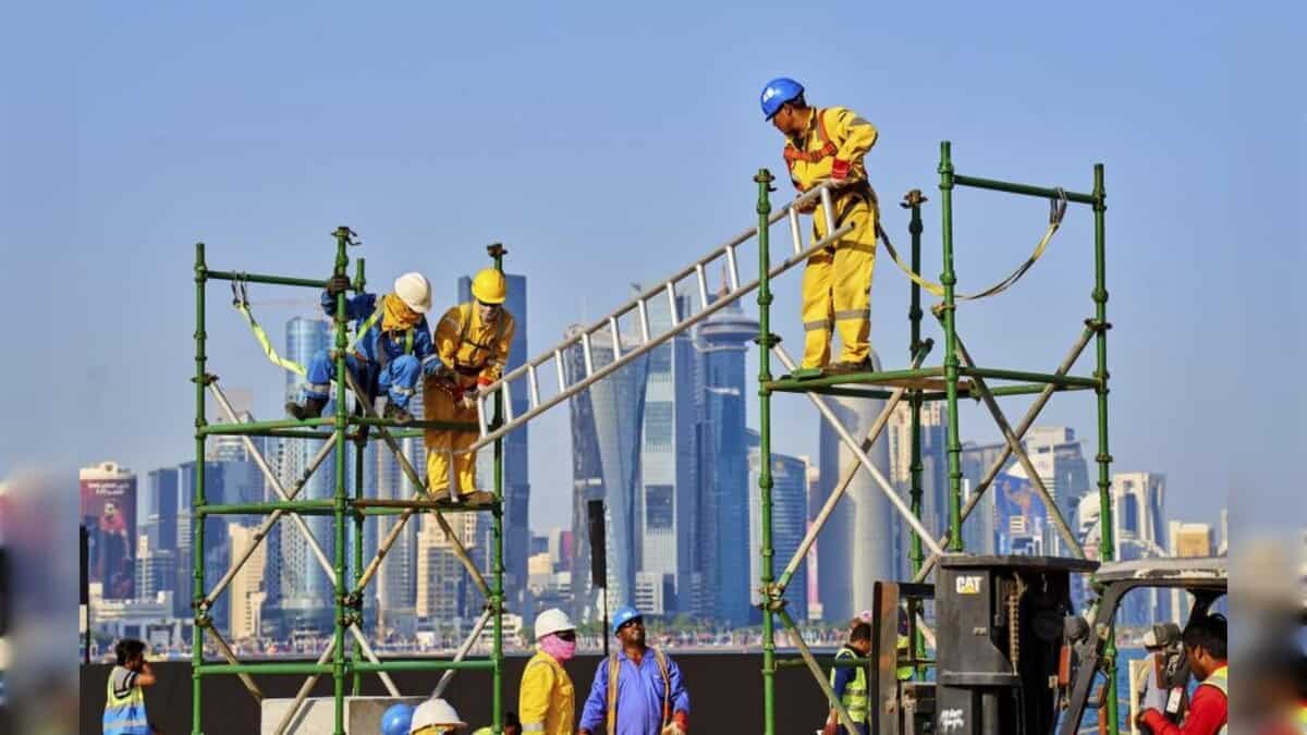 Migrant construction workers wearing safety gear and working on scaffolding with Gulf city skyscrapers in the background.