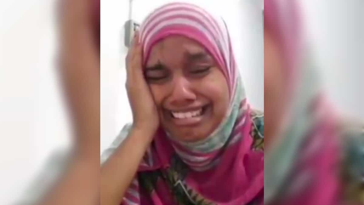 A woman wearing a pink and striped hijab is crying intensely, holding one hand to the side of her face. She appears distressed and emotional, with tears visible on her cheeks. The background is plain and indoor.