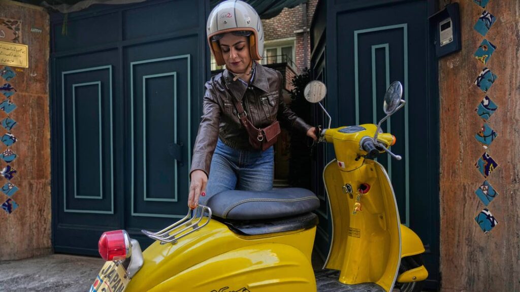 A woman wearing a helmet prepares her yellow scooter outside a building in Tehran, Iran.