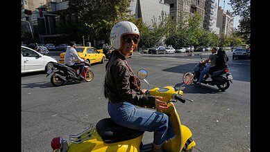 A woman rides a yellow scooter through a busy intersection in downtown Tehran, Iran.