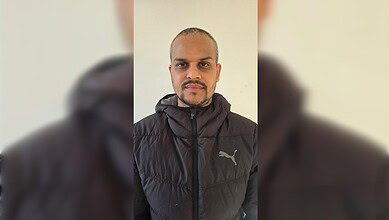 A man in a black winter jacket standing against a light-coloured wall.