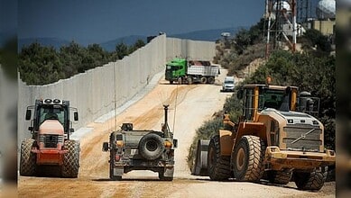 Construction vehicles and a military jeep operate along a concrete border wall near a wooded area.