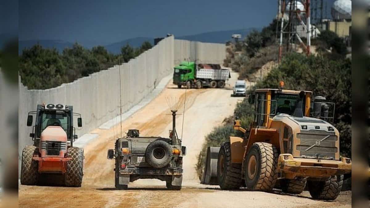 Construction vehicles and a military jeep operate along a concrete border wall near a wooded area.
