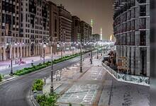 A quiet, well-lit street in Madinah at night with modern buildings and the Prophet’s Mosque minarets glowing in the distance.