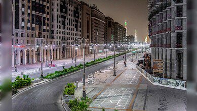 A quiet, well-lit street in Madinah at night with modern buildings and the Prophet’s Mosque minarets glowing in the distance.