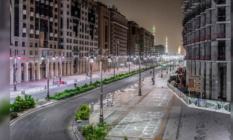 A quiet, well-lit street in Madinah at night with modern buildings and the Prophet’s Mosque minarets glowing in the distance.