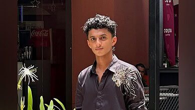 A young man wearing a dark shirt with a decorative design stands outdoors near plants and a cafe.