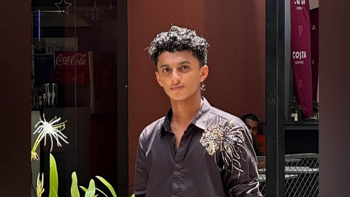 A young man wearing a dark shirt with a decorative design stands outdoors near plants and a cafe.