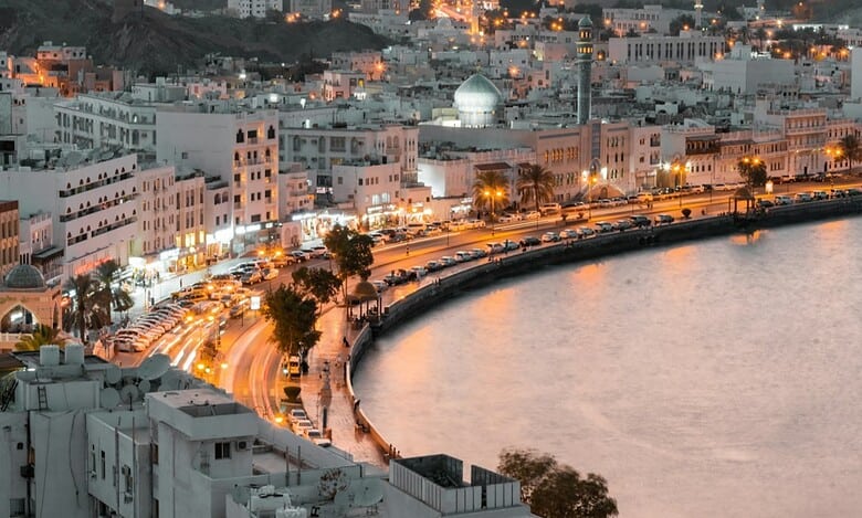A scenic evening view of Muttrah Corniche in Muscat, Oman, with city lights reflecting on the waterfront.