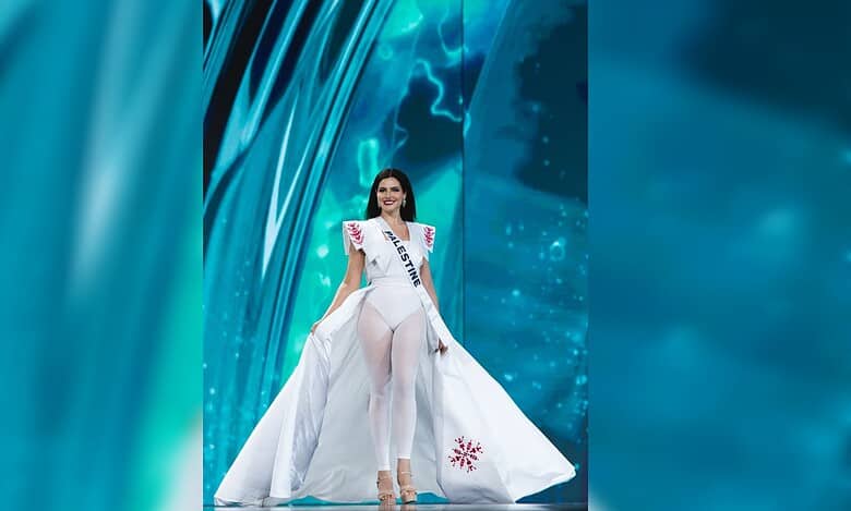 Miss Palestine Nadeen Ayoub walks on stage during the Miss Universe 2025 swimsuit round wearing a white embroidered cape and tights.