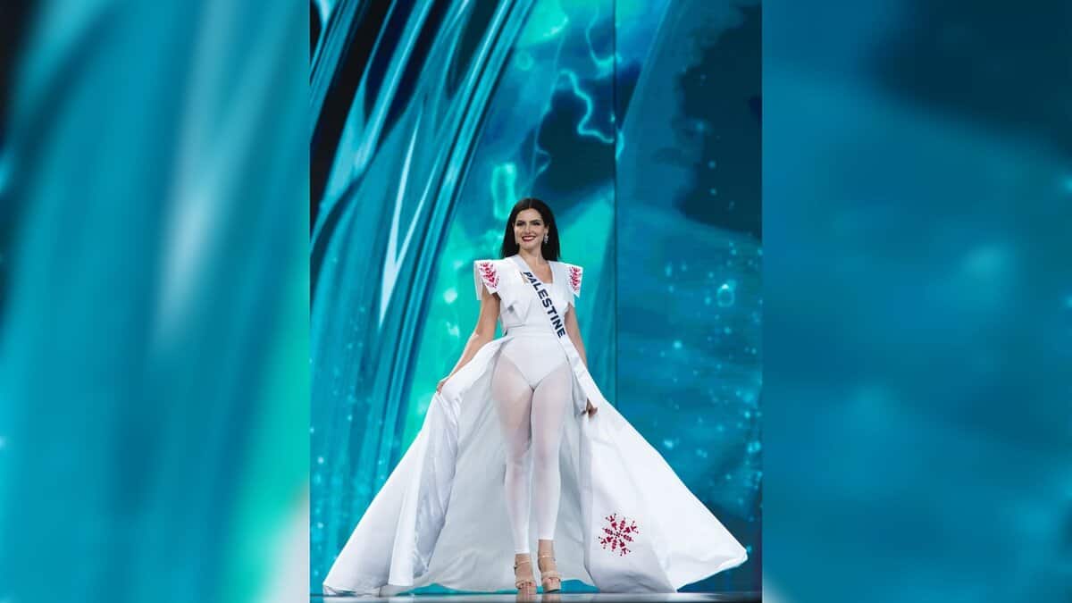 Miss Palestine Nadeen Ayoub walks on stage during the Miss Universe 2025 swimsuit round wearing a white embroidered cape and tights.