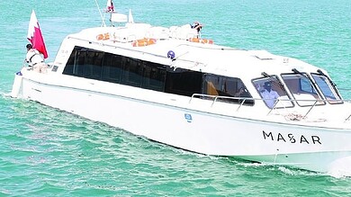 A white passenger ferry named MASAR, carrying the Qatari flag, sails through turquoise waters at Al Ruwais Port in Qatar.