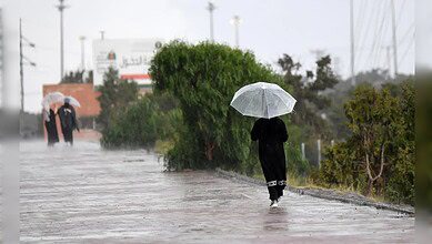 A woman walks along a rain-soaked pathway holding a transparent umbrella, while two others with umbrellas stand in the background during heavy rainfall.