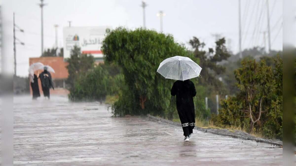 A woman walks along a rain-soaked pathway holding a transparent umbrella, while two others with umbrellas stand in the background during heavy rainfall.
