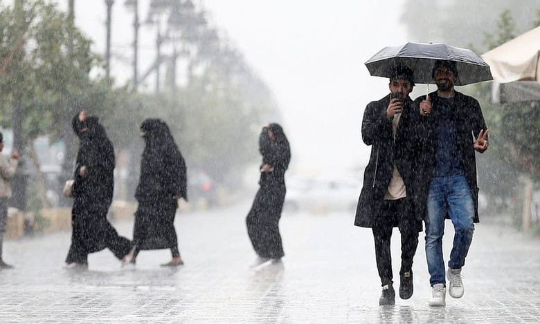 People walk through heavy rain in Saudi Arabia, some using umbrellas while others cross the street in wet conditions.