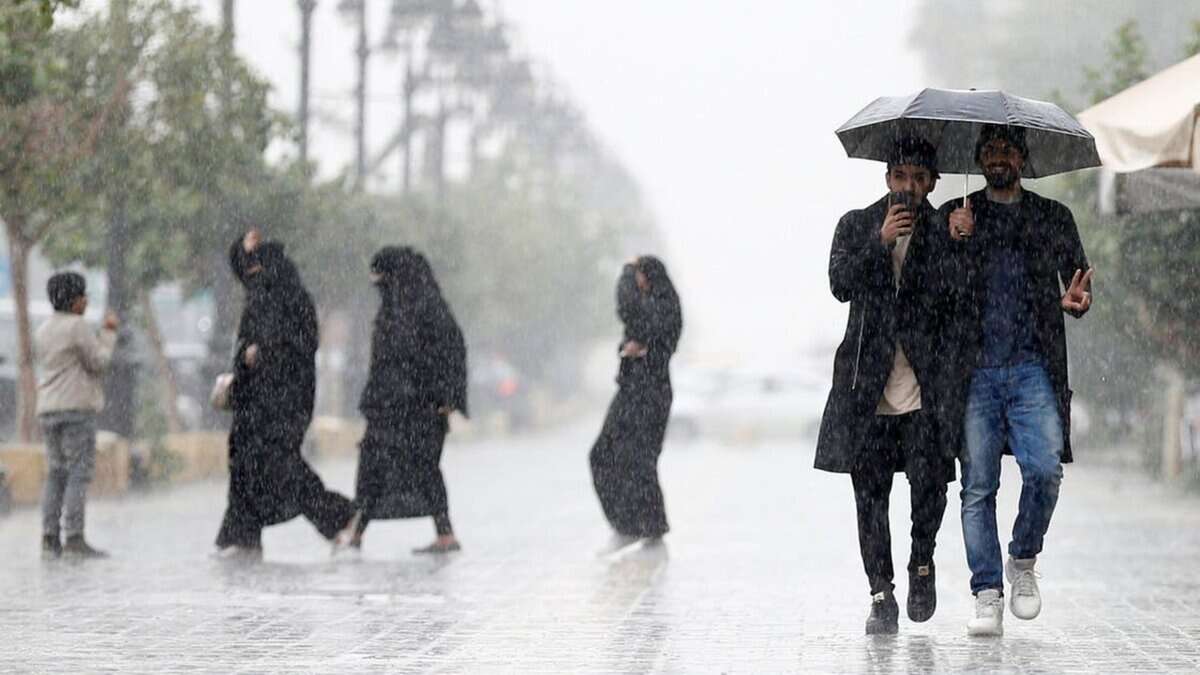People walk through heavy rain in Saudi Arabia, some using umbrellas while others cross the street in wet conditions.