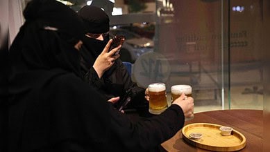 Two Saudi women in niqab sit at a cafe table in Riyadh, holding glasses of alcohol-free draft beer while one of them takes a photo on her phone.