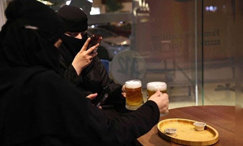 Two Saudi women in niqab sit at a cafe table in Riyadh, holding glasses of alcohol-free draft beer while one of them takes a photo on her phone.